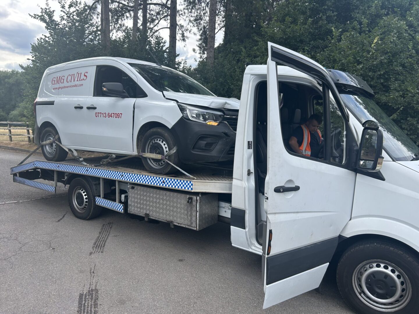A damaged white van being towed on a flatbed truck.